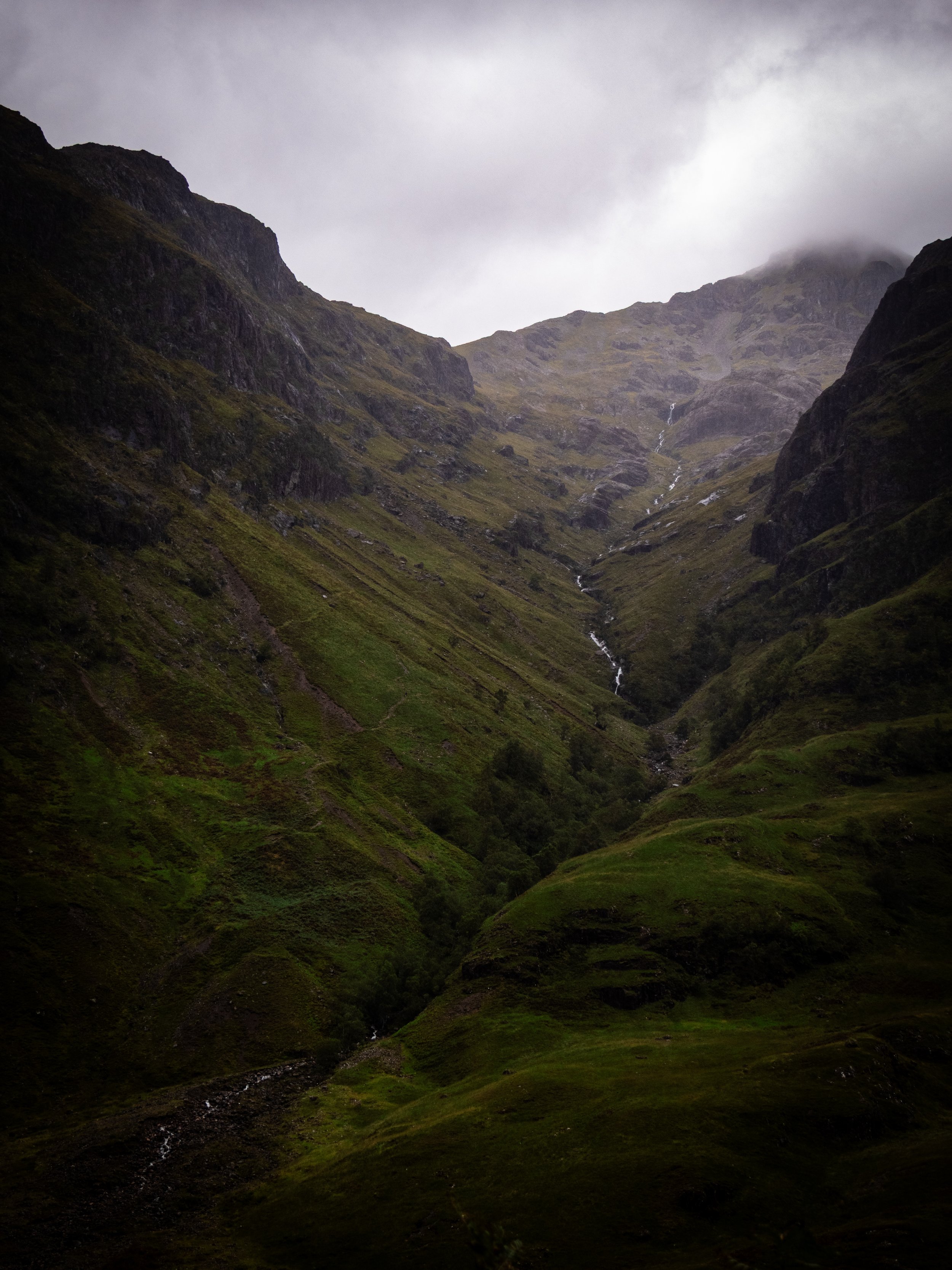 A misty mountain valley with steep green slopes and small waterfalls.