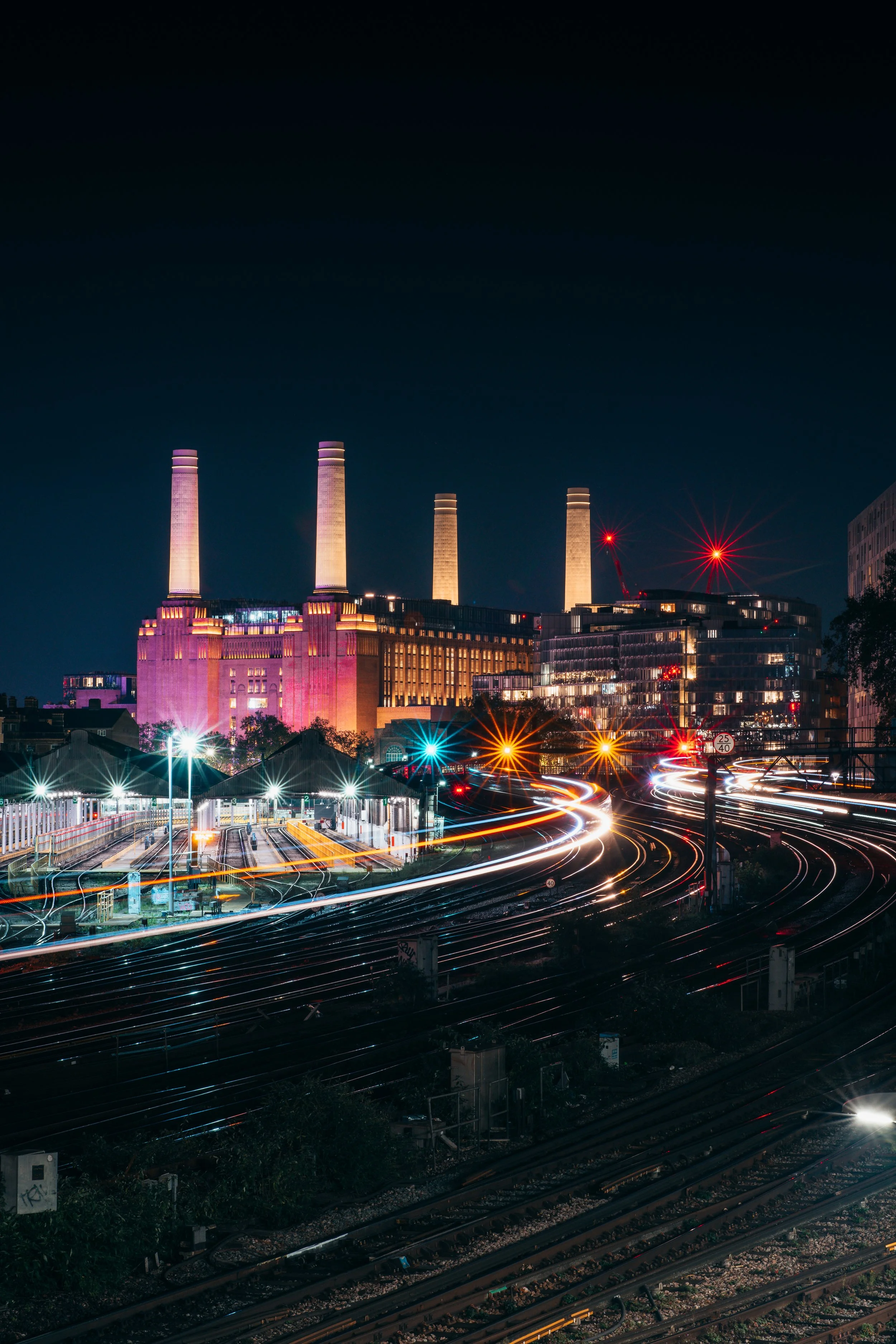 Nighttime cityscape of a power plant with four tall smokestacks illuminated, surrounded by illuminated railway tracks and light trails from moving vehicles.
