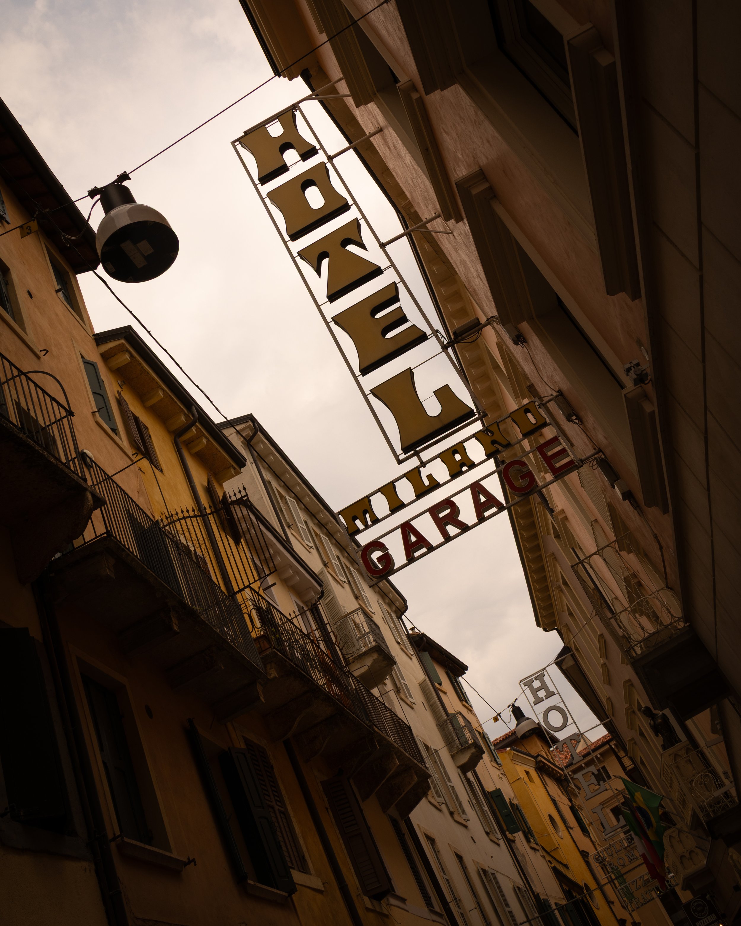 View of narrow street with old European-style buildings, hanging signs for a hotel, garage, and other businesses, with cloudy sky above.