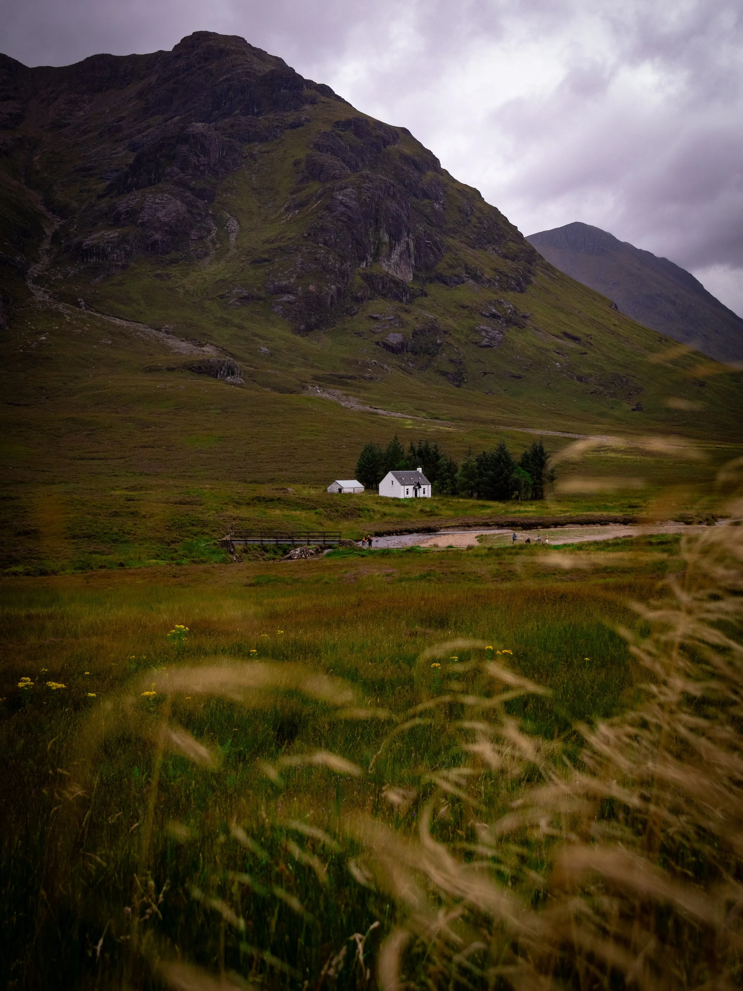 A rural landscape with green grassy fields, a small river, and three white houses surrounded by trees at the base of tall, rugged mountains under a cloudy sky.
