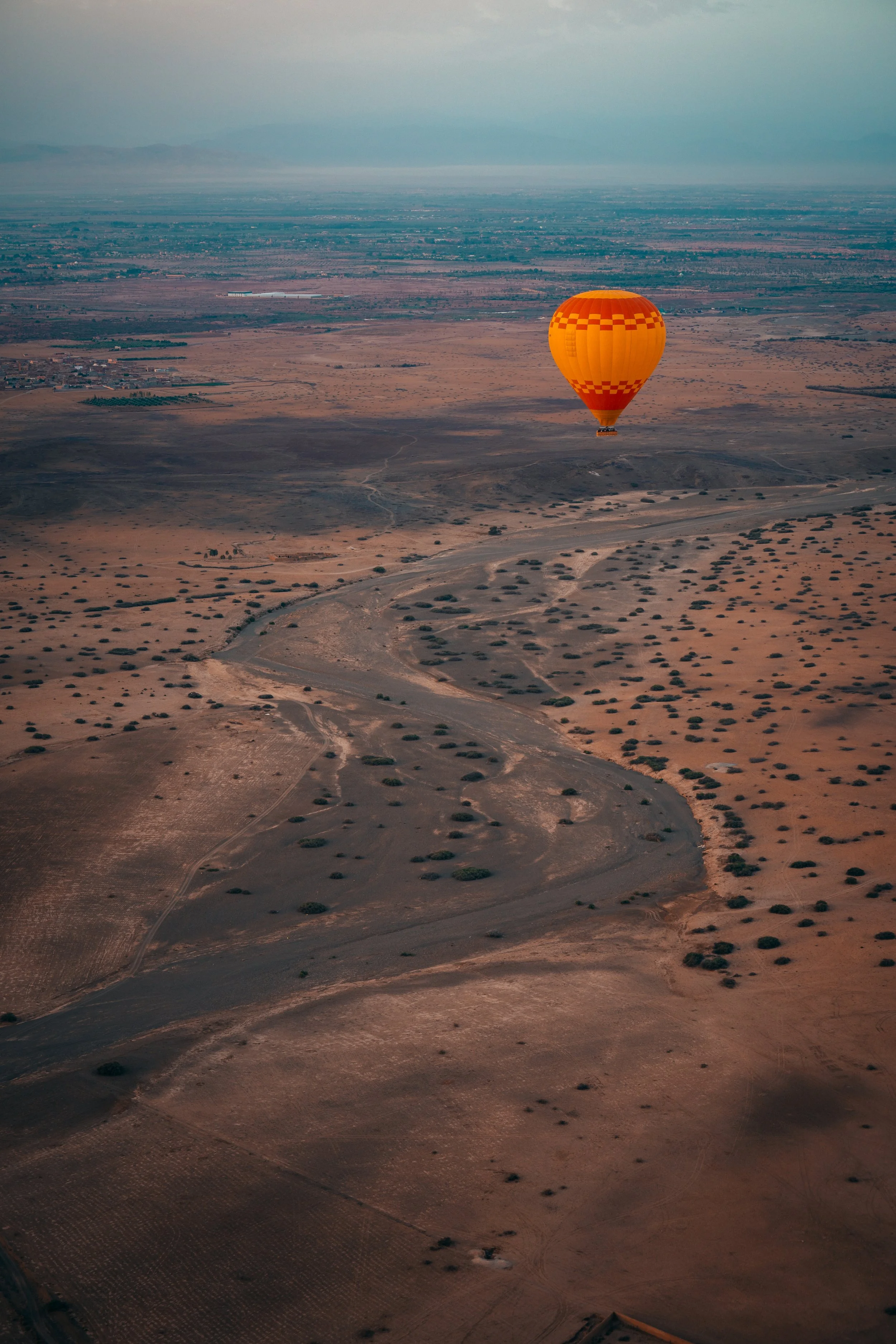 A hot air balloon flying over a vast, arid landscape with sparse vegetation and dirt paths during sunset or sunrise.