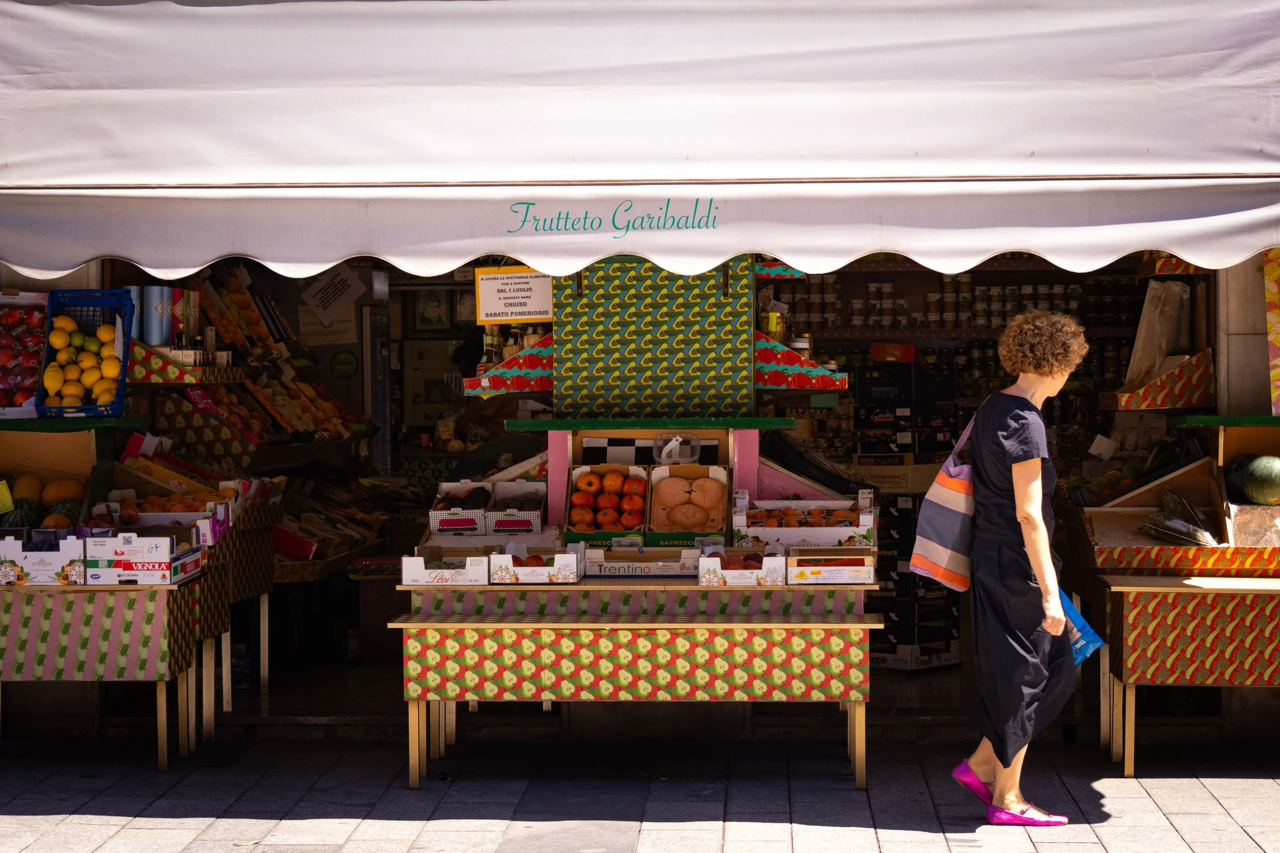 A woman with curly hair wearing a black dress, pink shoes, and carrying a striped bag, walking past a fruit stand with colorful fruit displays and a white canopy.