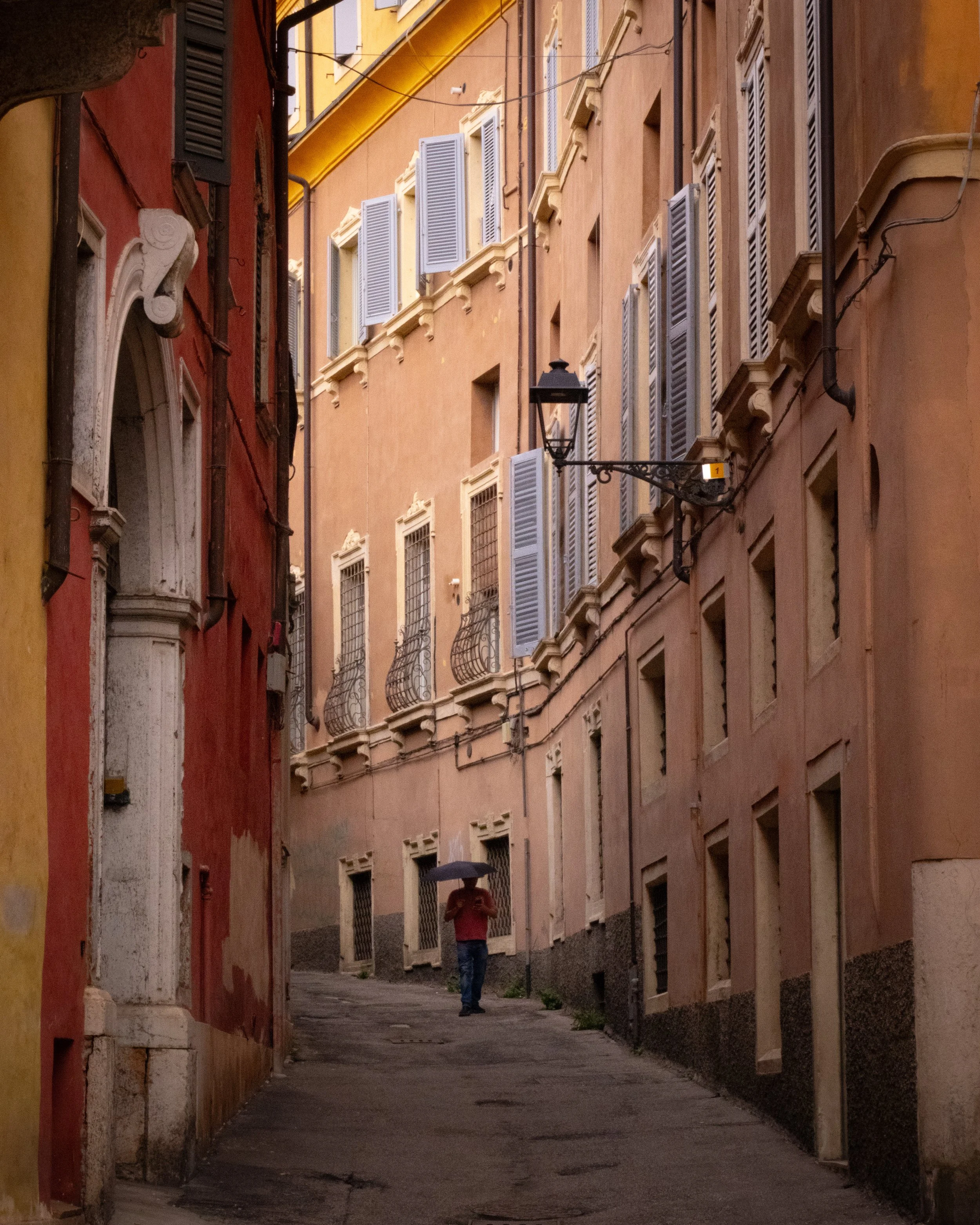 A narrow street in a European city, with pastel-colored buildings and small balconies, a person walking with an umbrella.