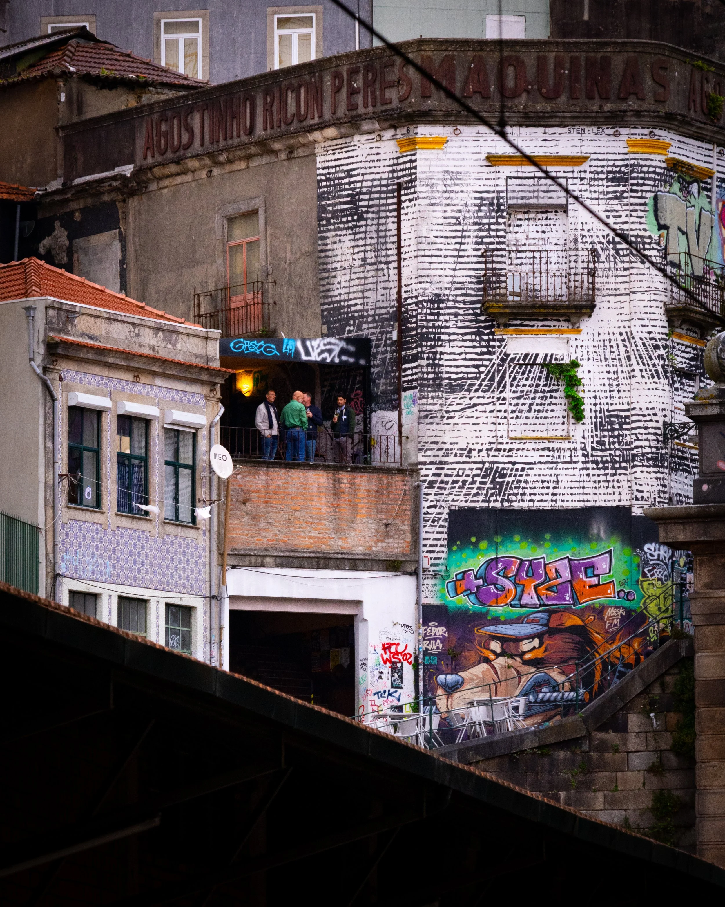 A multi-story building with graffiti art, including a colorful mouse character and the word 'SHOE,' and a group of people conversing on a balcony.