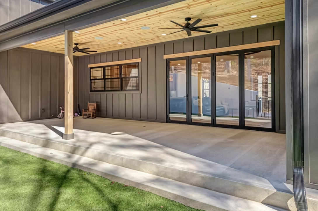 A modern backyard patio with a concrete floor, sliding glass doors, dark exterior walls, and a wooden ceiling with two ceiling fans.