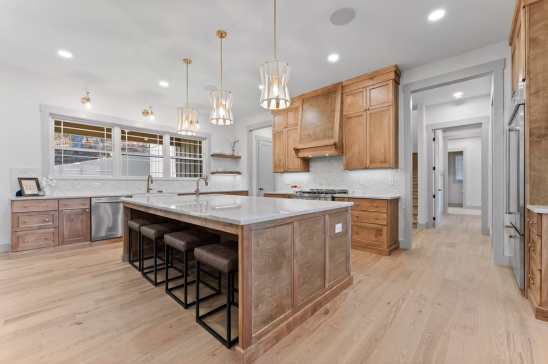 Modern kitchen with wooden cabinetry, marble island with four stools, large windows, and pendant lighting.