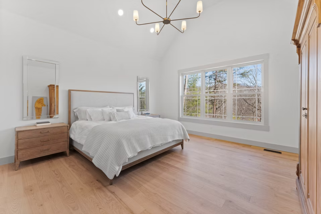Bright bedroom with large window, white walls, wooden furniture, bed with white bedding, and a modern chandelier
