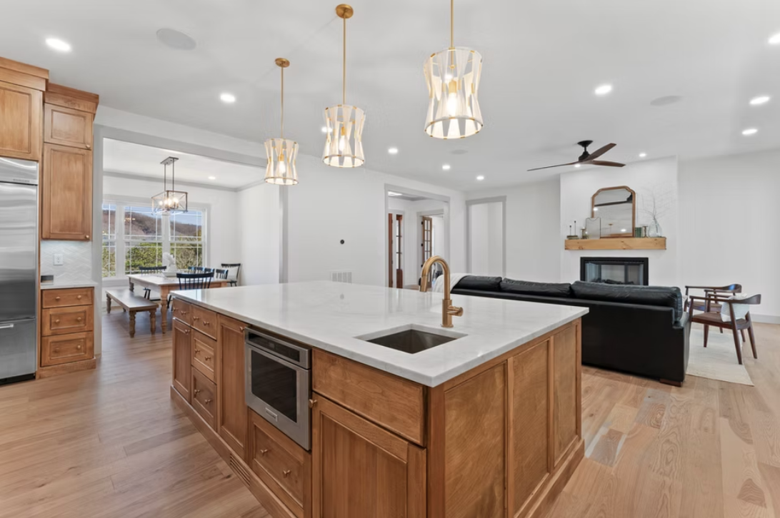 Open-concept kitchen with wooden cabinetry, white marble island, three pendant lights, and a living area with a sofa, fireplace, and ceiling fan.