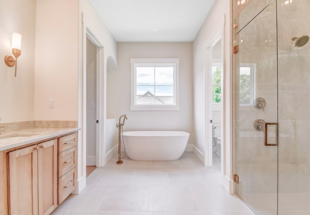 Bright bathroom with a freestanding bathtub beneath a window, glass-enclosed shower on the right, and a wooden vanity with a marble countertop on the left.