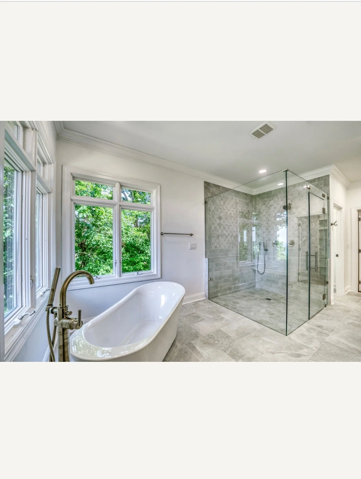 Modern bathroom featuring a freestanding bathtub with a brass faucet and a large glass-enclosed shower with gray tile walls and floors, and multiple windows with greenery outside.