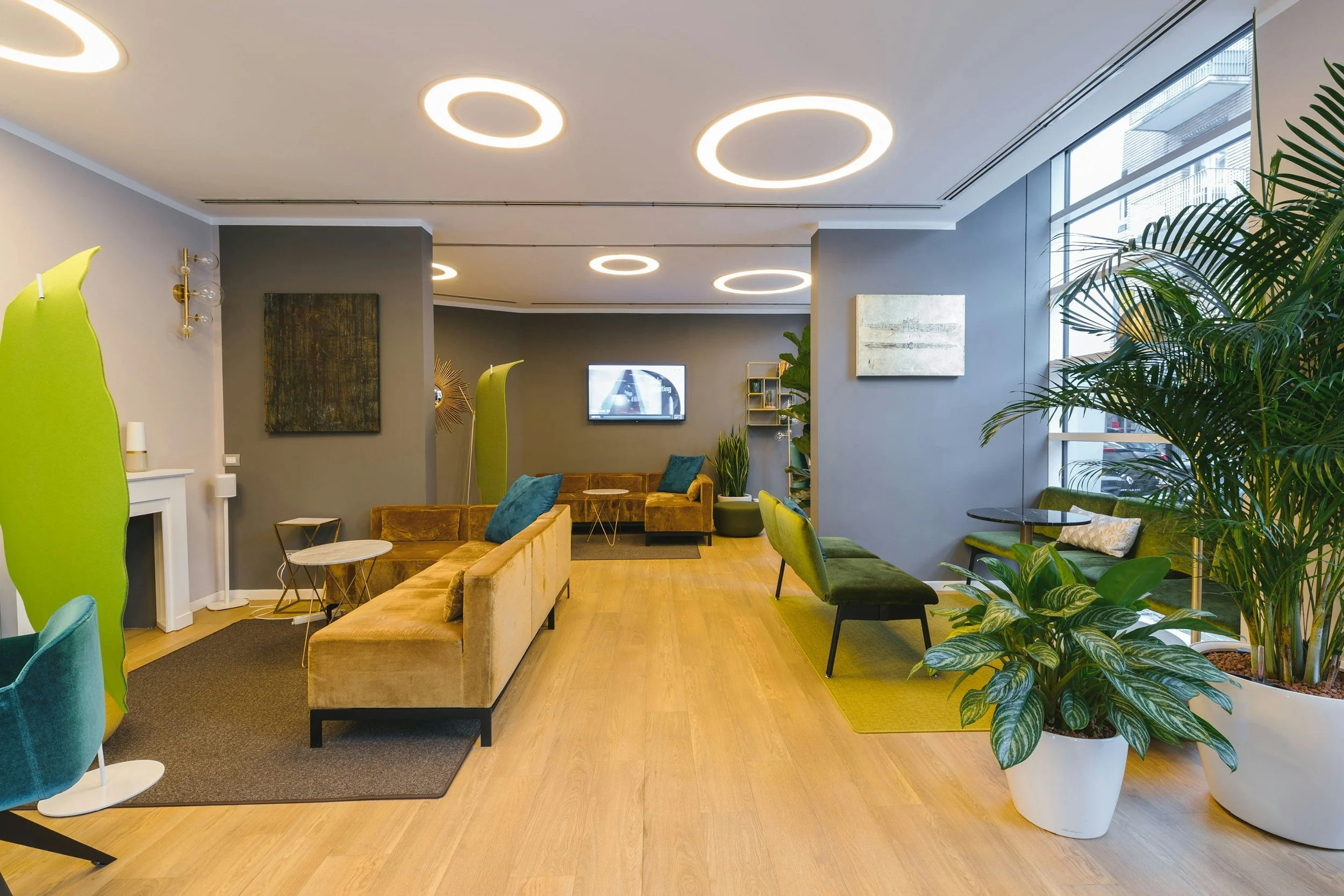 Modern lobby with green and mustard-colored sofas, green chairs, large potted plants, wooden flooring, and contemporary lighting fixtures.