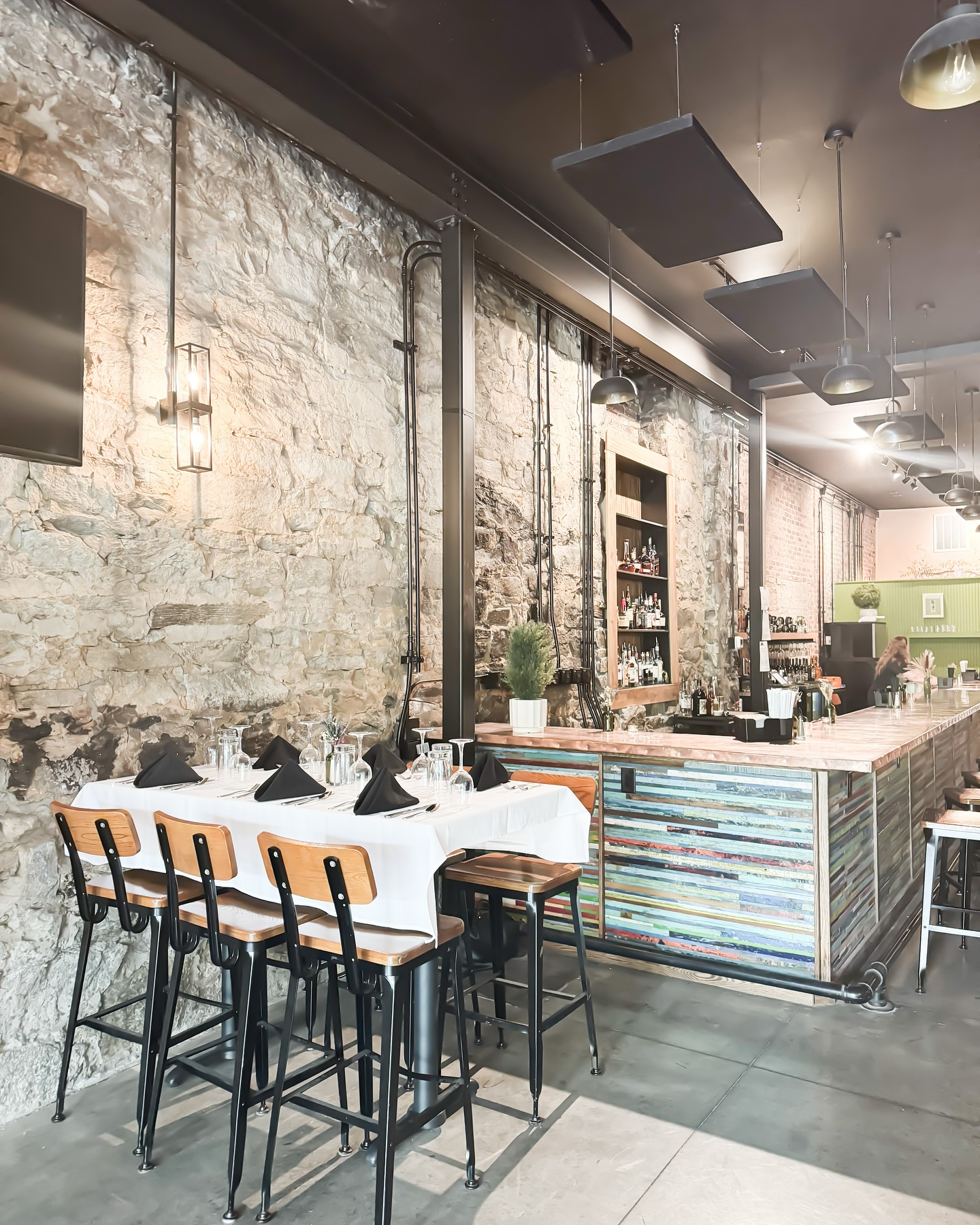 Interior of a modern restaurant with a stone wall, a white dining table set with black napkins and glassware, and a colorful bar counter.