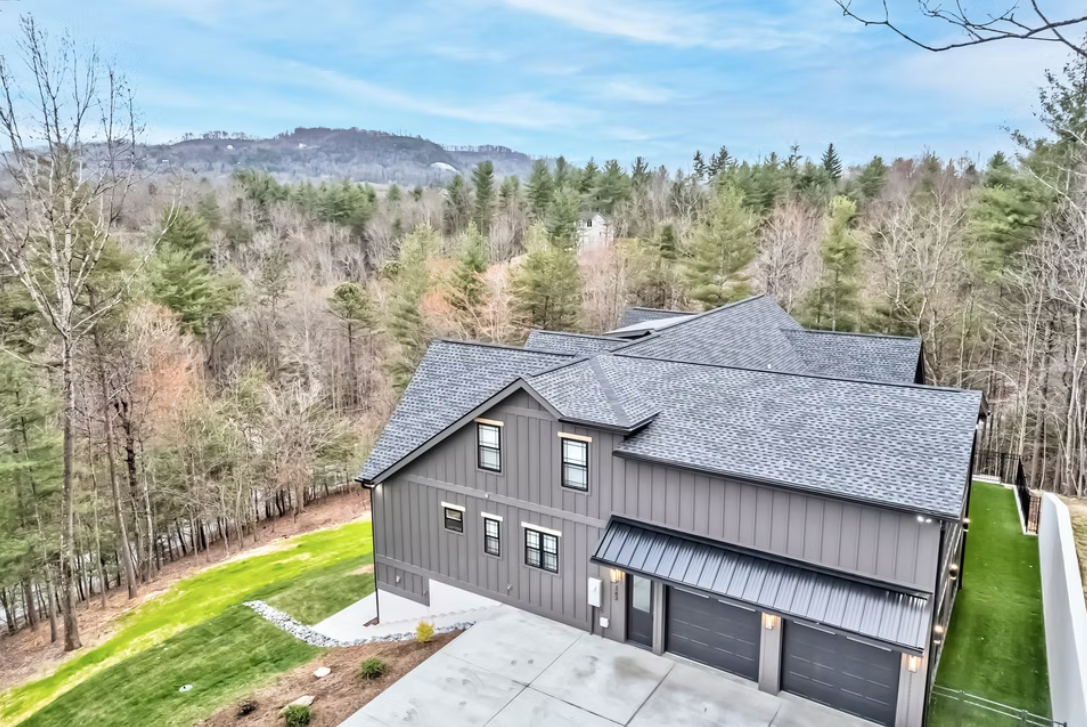 A modern gray house with a black roof and a black garage door, surrounded by a green lawn and trees in a wooded area.