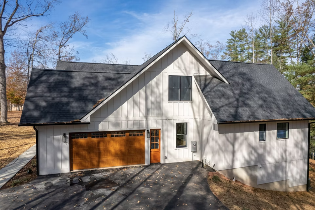 A modern two-story house with white exterior siding, a black shingled roof, and a wooden garage door, situated among leafless trees on a clear day.