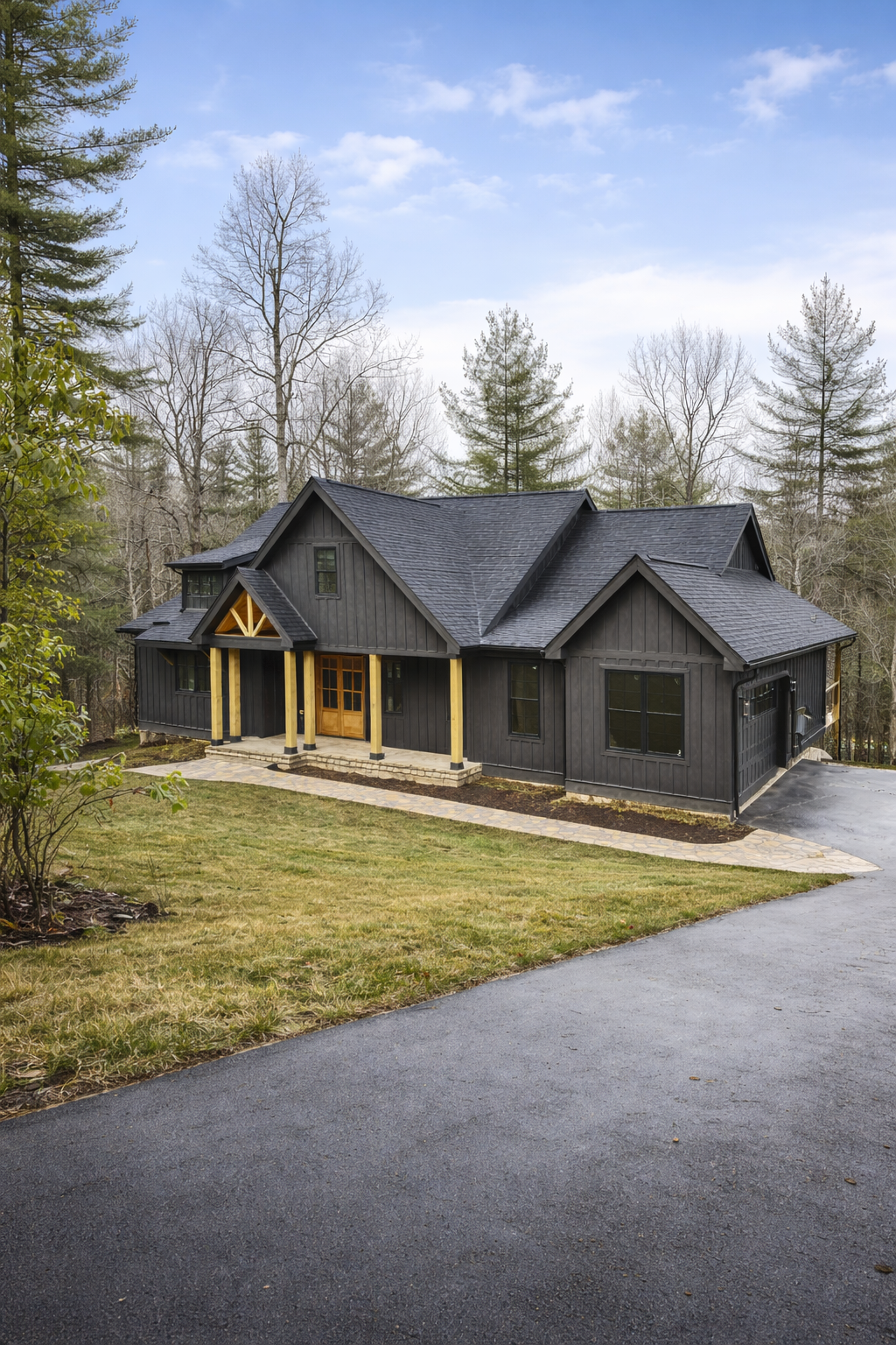 A black wooden house with a gray shingled roof surrounded by trees, with a stone pathway and driveway in front.