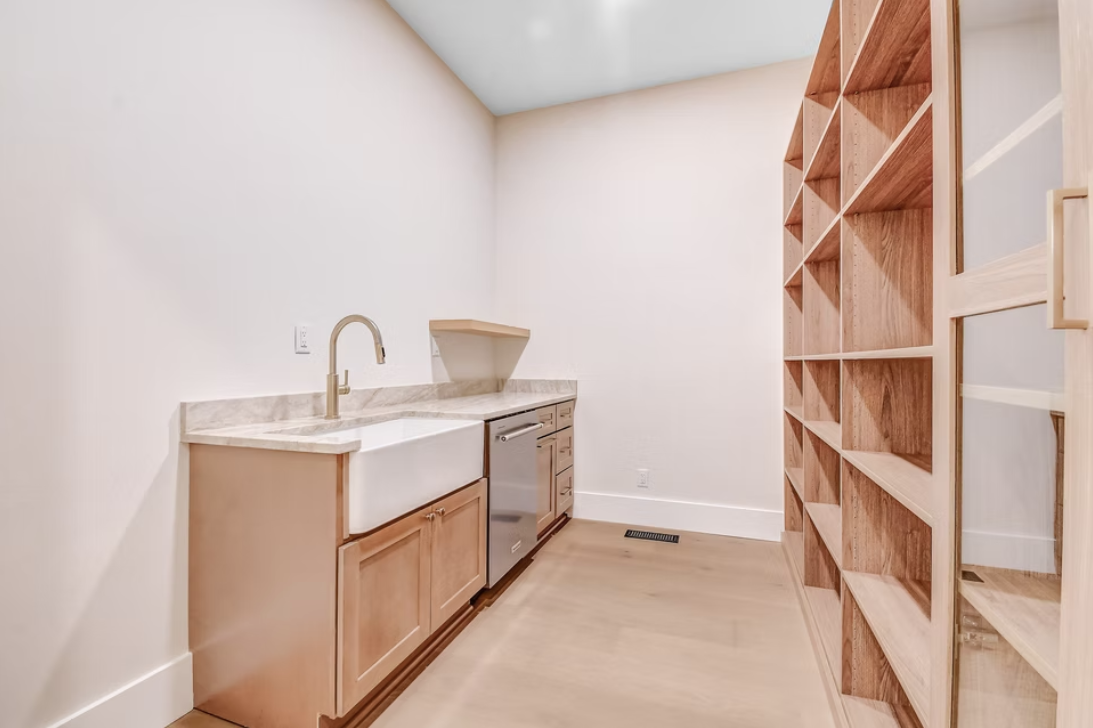 Empty kitchenette with a farmhouse sink, marble countertop, and wooden cabinets on the left; open wooden shelving on the right; light-colored walls and flooring.