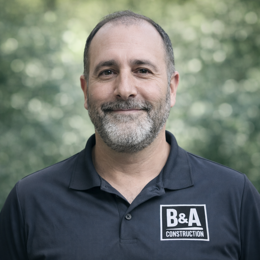 Portrait of a middle-aged man with a beard, wearing a black polo shirt with a B&A Construction logo, standing outdoors with blurred green foliage in the background.