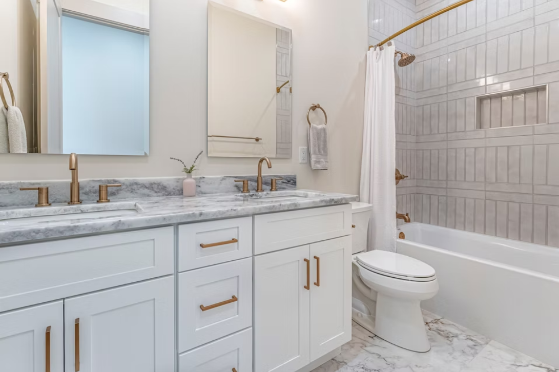 Modern bathroom with white cabinetry, marble countertops, gold fixtures, a bathtub with a tiled wall, and a toilet.
