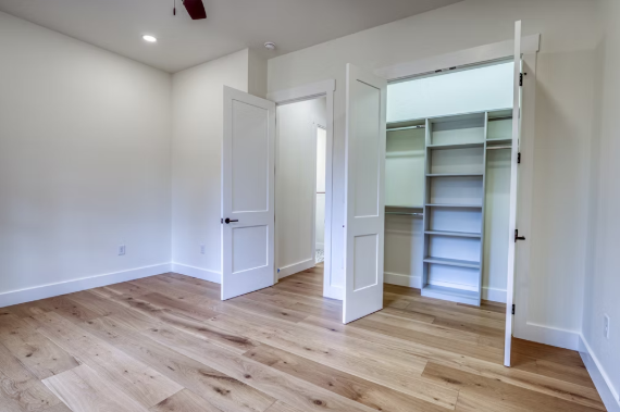 Empty room with hardwood floors, white walls, open closet with shelves and a door, and a ceiling fan.