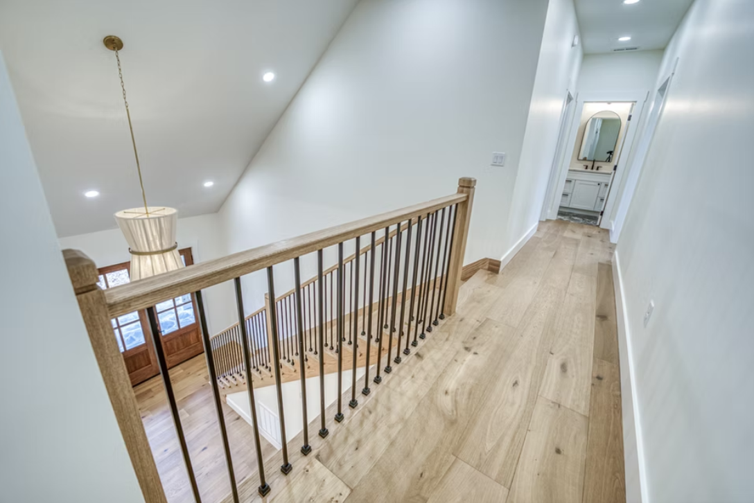 Interior view of a second-floor hallway with wooden flooring, white walls, and a wooden and metal railing overlooking a staircase. There is a hanging light fixture and a bathroom at the end of the hallway.