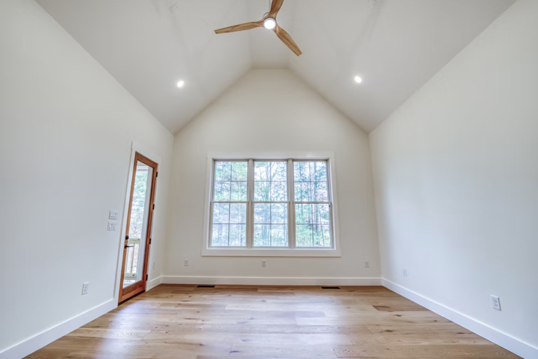 Empty room with wood flooring, white walls, a large window, a door leading to a balcony, and a ceiling fan with wooden blades.