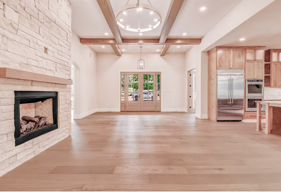Spacious open living room with wooden floors, a large stone fireplace on the left, wooden beams on the ceiling, and front double doors with glass panels. The right side features a kitchen with light wood cabinets and stainless steel appliances.