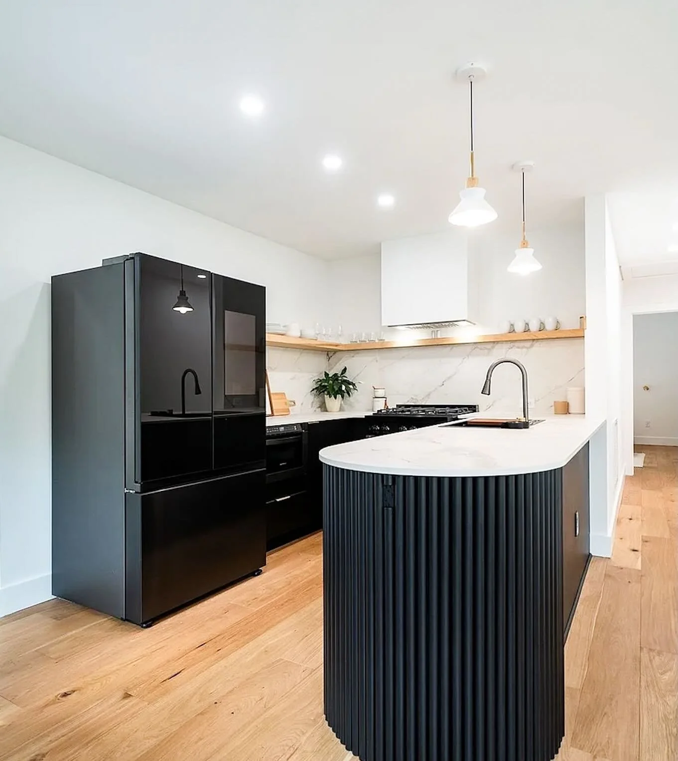 Modern kitchen with black refrigerator, white marble countertop, black cabinetry, and wooden accents, illuminated by pendant and recessed lighting.