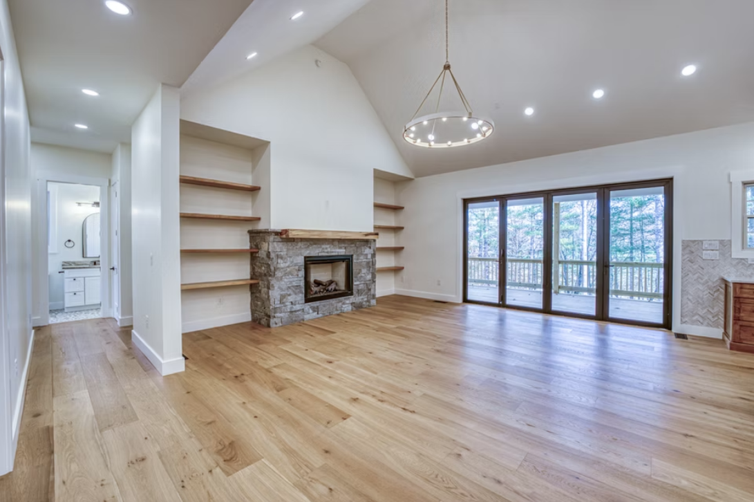 Empty living room with hardwood floors, a stone fireplace, built-in shelves, large sliding glass doors leading to a deck, and a modern chandelier hanging from a high ceiling.