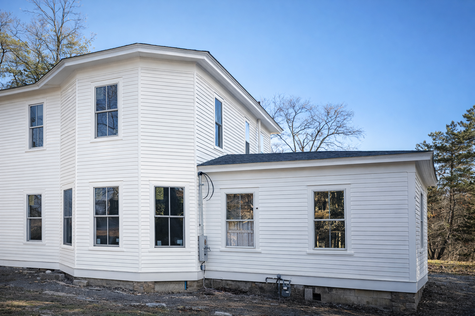 Front of a white, multi-story house with vinyl siding and several windows, set against a clear blue sky and surrounded by leafless trees.