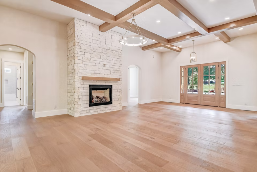 Empty living room with hardwood floor, stone fireplace, large windows, and wooden ceiling beams.