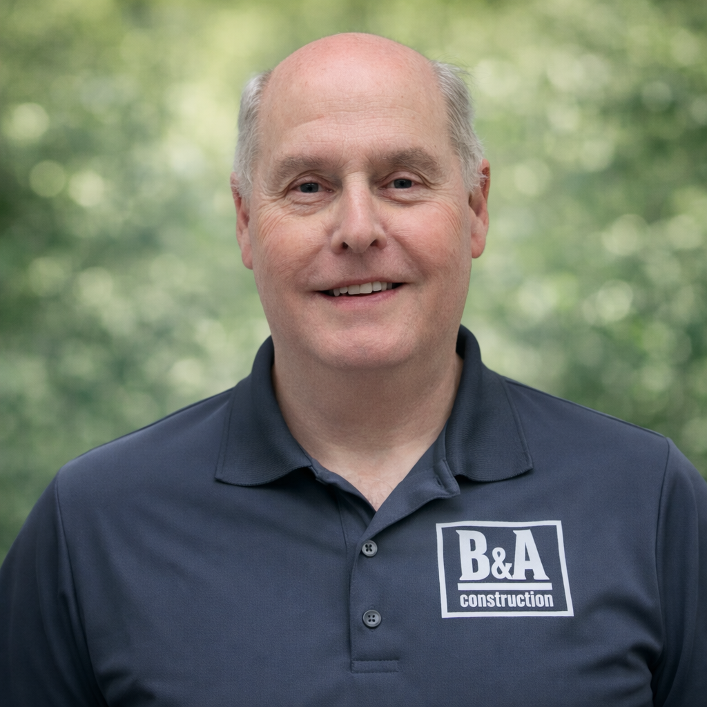A man with a bald head smiling at the camera, wearing a navy blue polo shirt with a 'B&A Construction' logo, outdoors with a blurred green leafy background.