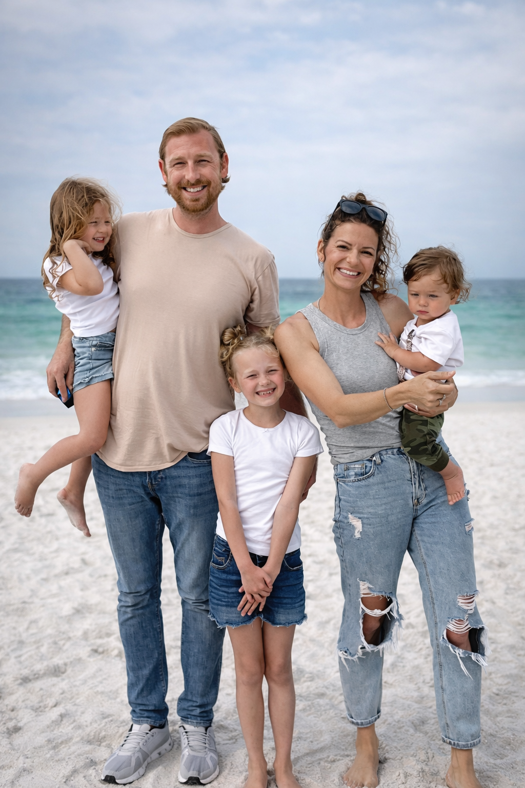 A smiling family of six on a beach, with the ocean in the background. The parents are holding two young children, and two older children stand in front of them.
