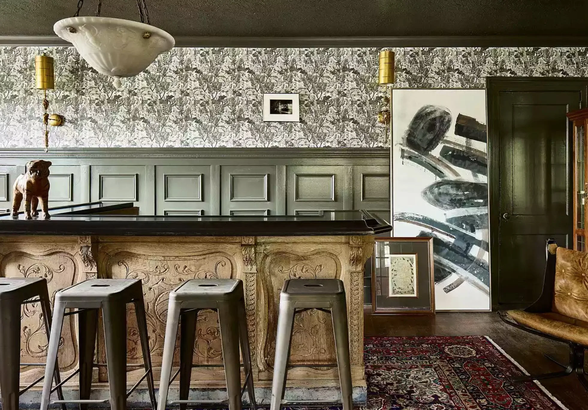 Interior of a vintage-style room with a bar counter, three black stools, framed artwork, patterned wallpaper, chandelier, and a leather chair.
