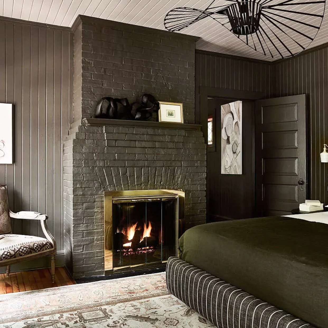 Living room with a lit fireplace, dark wood paneled walls, a bed with a dark green cover, a chair with patterned upholstery, and decorative items on the wall and shelves.