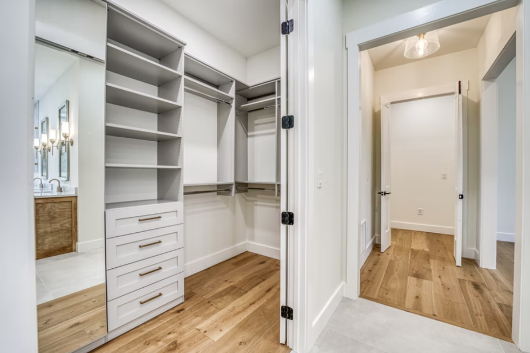 Empty walk-in closet with white built-in shelves, drawers, and hanging space, with hardwood flooring and an adjacent bathroom with a mirror and bathroom fixtures visible.