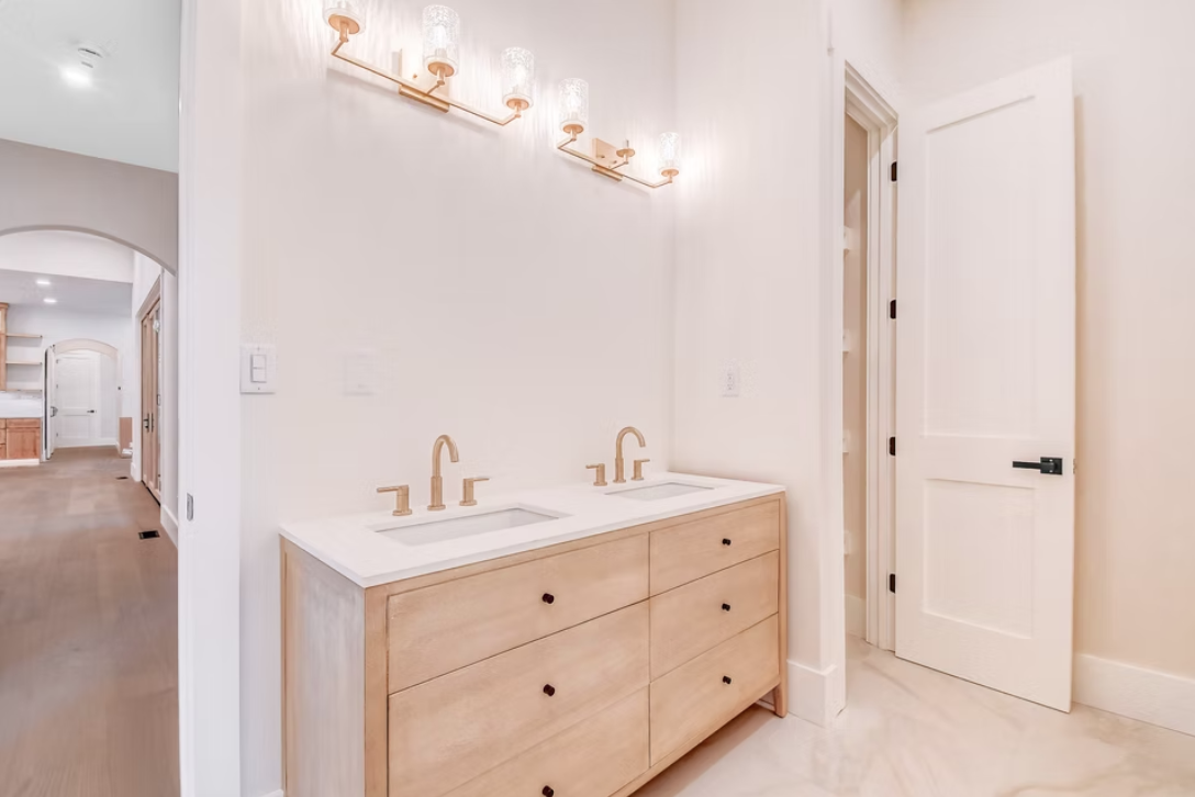 Modern bathroom with dual sinks, wooden vanity, gold fixtures, and a wall-mounted light fixture above.
