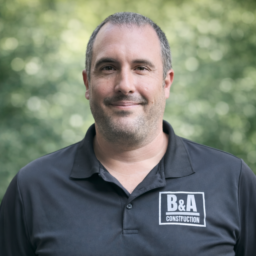 A man smiling outdoors, wearing a black polo shirt with a B&A Construction logo, with a blurred green natural background.