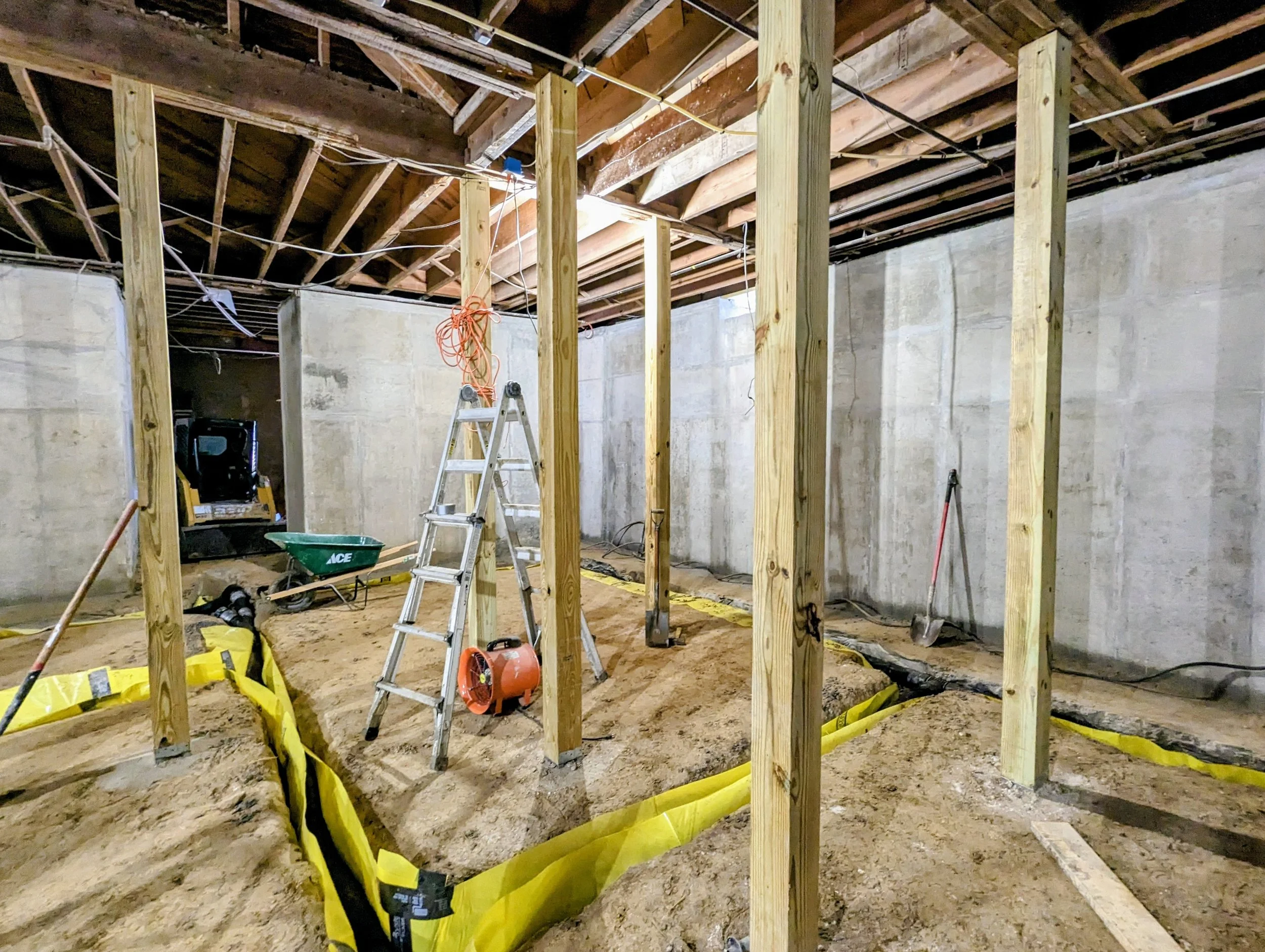 Construction site of a basement or interior space with wooden framing, concrete walls, and exposed ceiling beams, with construction tools and materials including a ladder, wheelbarrow, shovel, and yellow safety barriers.