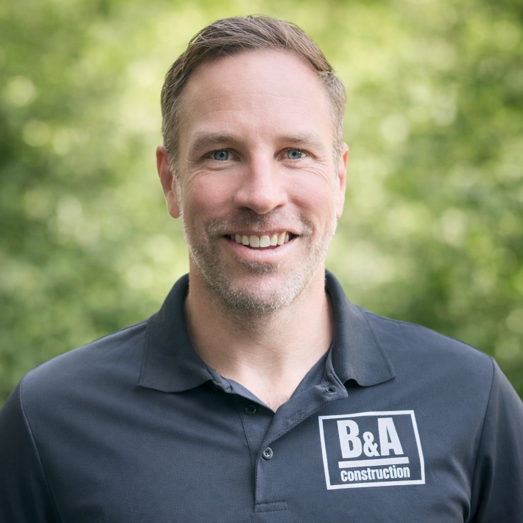 A man smiling outdoors with a blurred green background, wearing a dark blue polo shirt with a "B&A construction" logo.