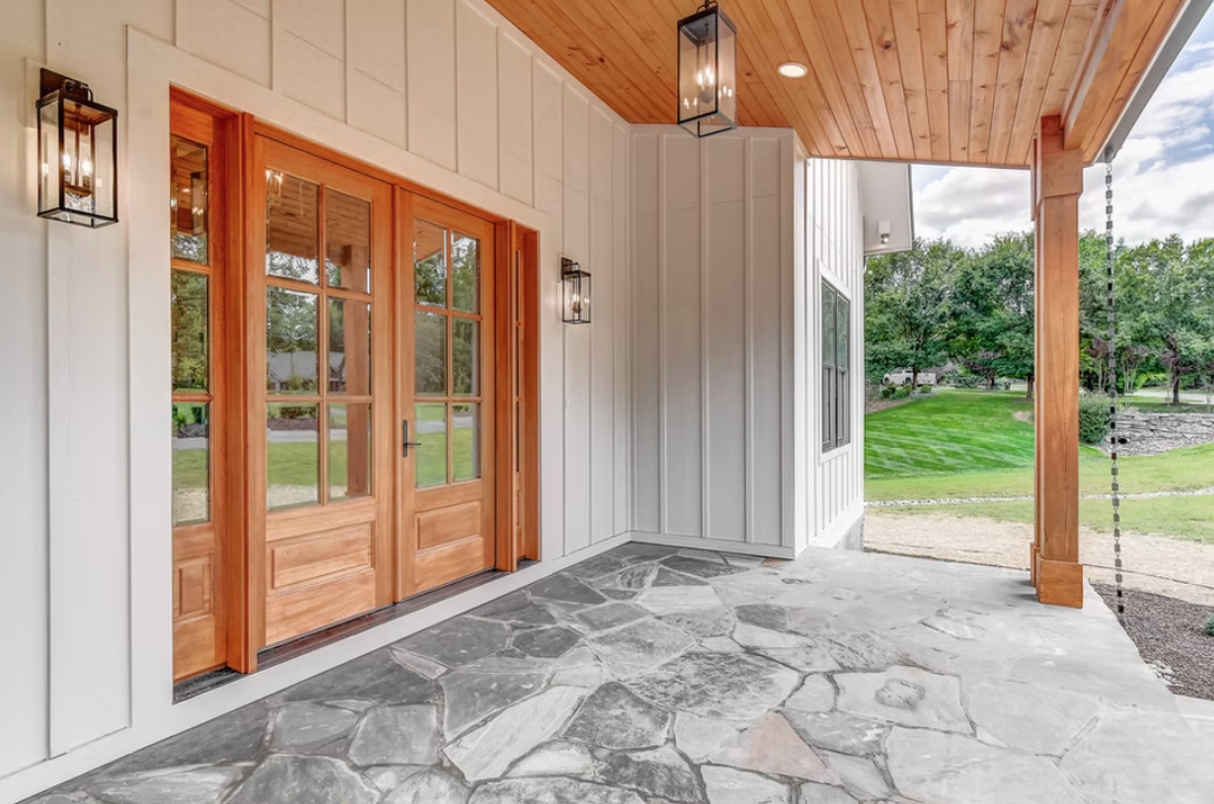 Front porch with stone flooring, wooden double doors with glass panels, black lantern-style wall sconces, and a wooden ceiling. Green lawn and trees are visible outside.
