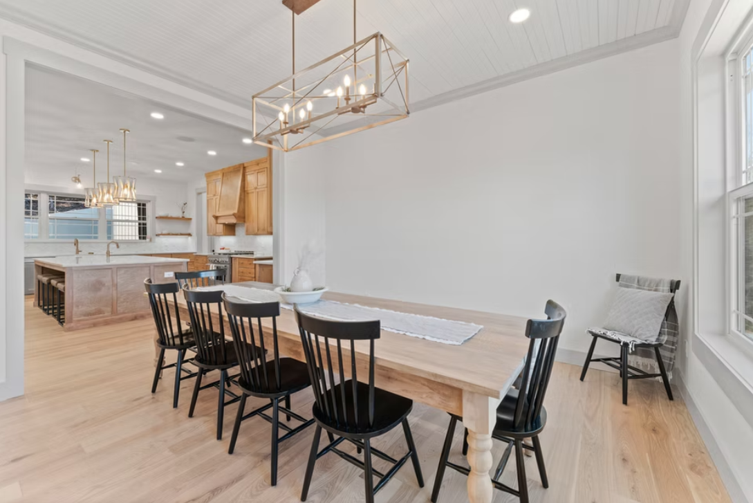 Dining area with a wooden table, six black chairs, a decorative bowl, and a gold geometric chandelier. Adjacent kitchen with wooden cabinets and an island.