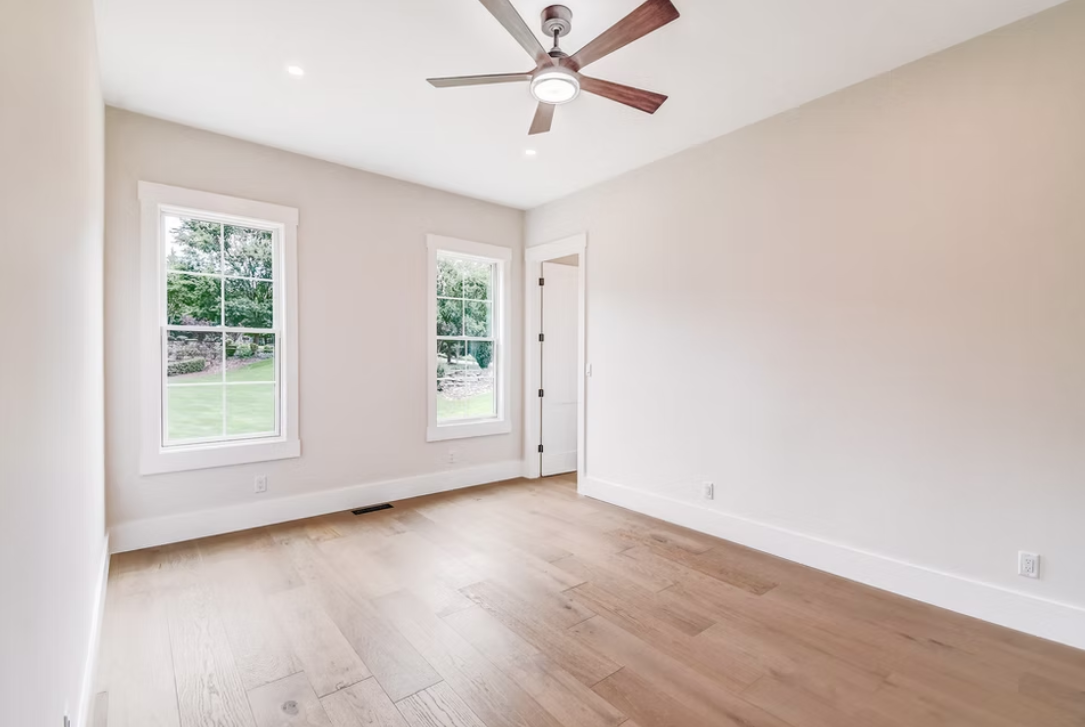 Empty room with white walls, light wood flooring, two windows, a white door, a ceiling fan with brown blades, and a ceiling light, with a view of a green lawn outside.