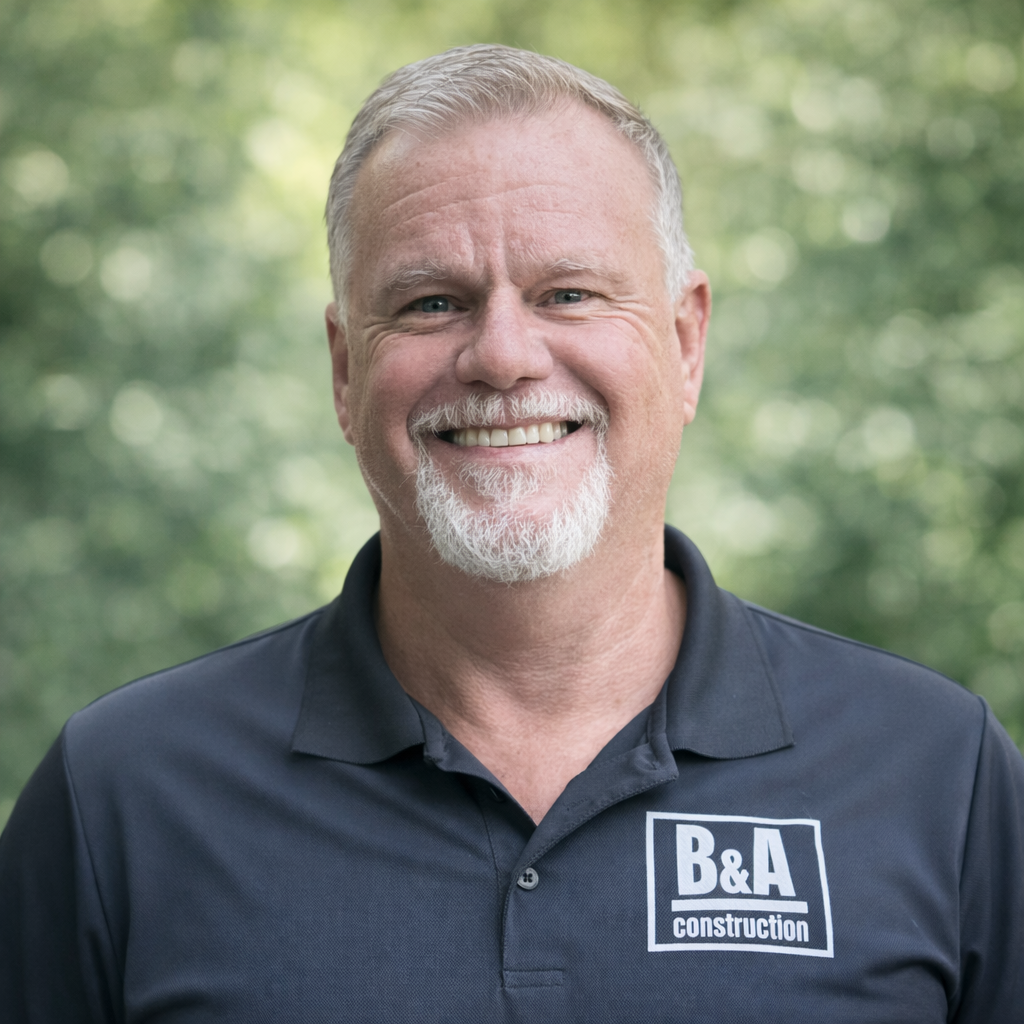 Smiling man with short gray hair, beard, and mustache wearing a black polo shirt with 'B&A Construction' logo, outdoors with blurred greenery background.