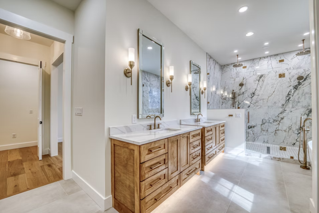 Modern bathroom with double vanity sinks, gold fixtures, mirrors, wall sconces, and a marble walk-in shower with gold accents.