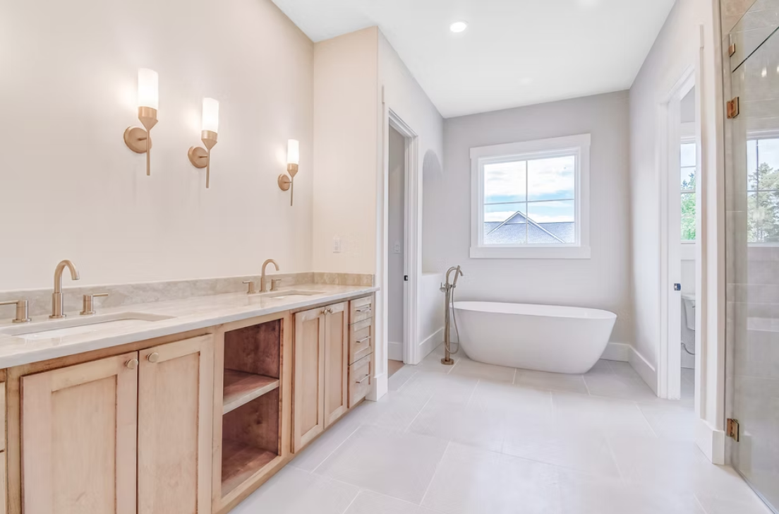 Modern bathroom with double vanity, wall-mounted lights, and a freestanding bathtub by a window.