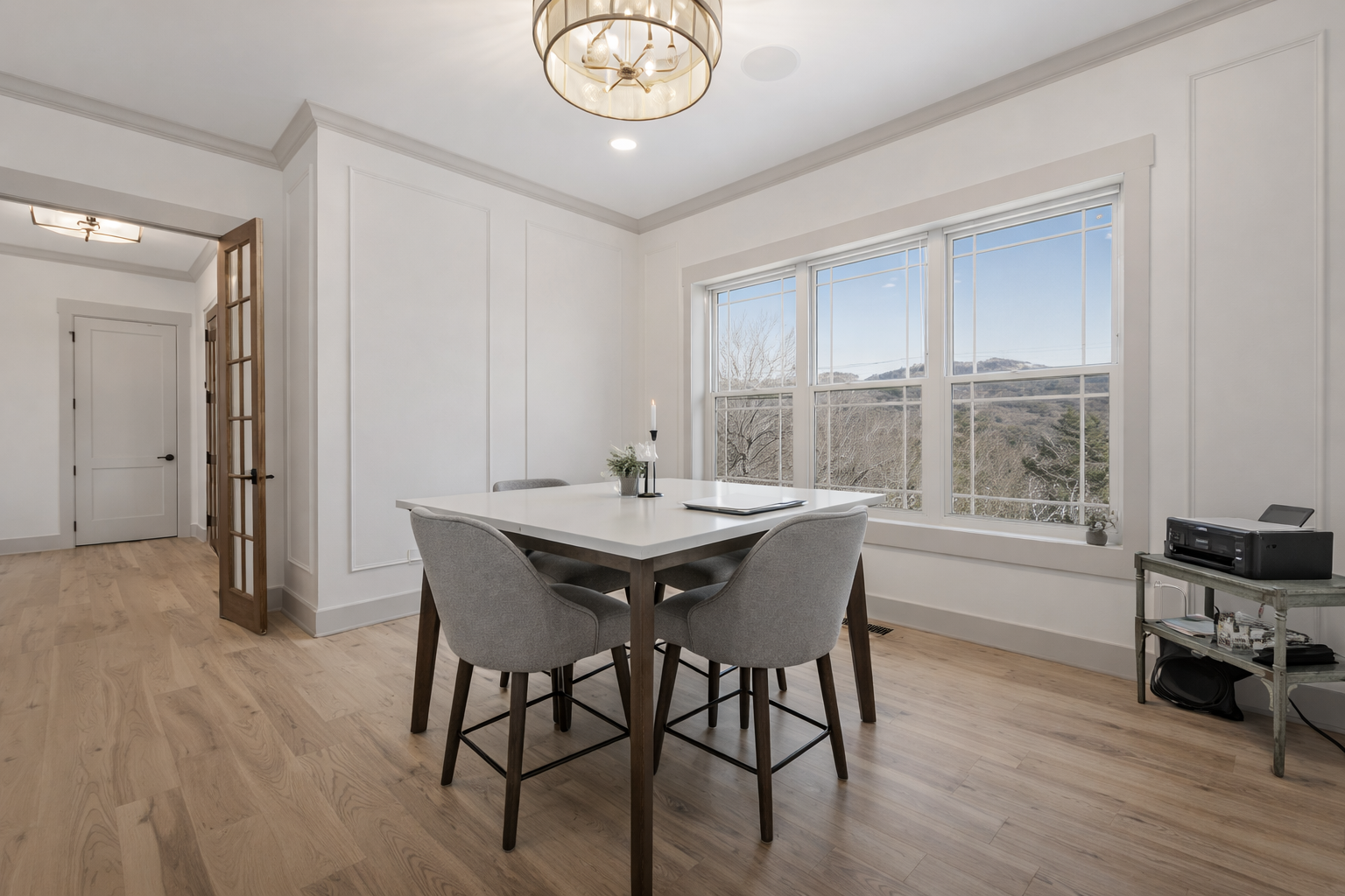 Dining room with white table, four gray chairs, large window with mountains view, wooden floor, and minimal decor.
