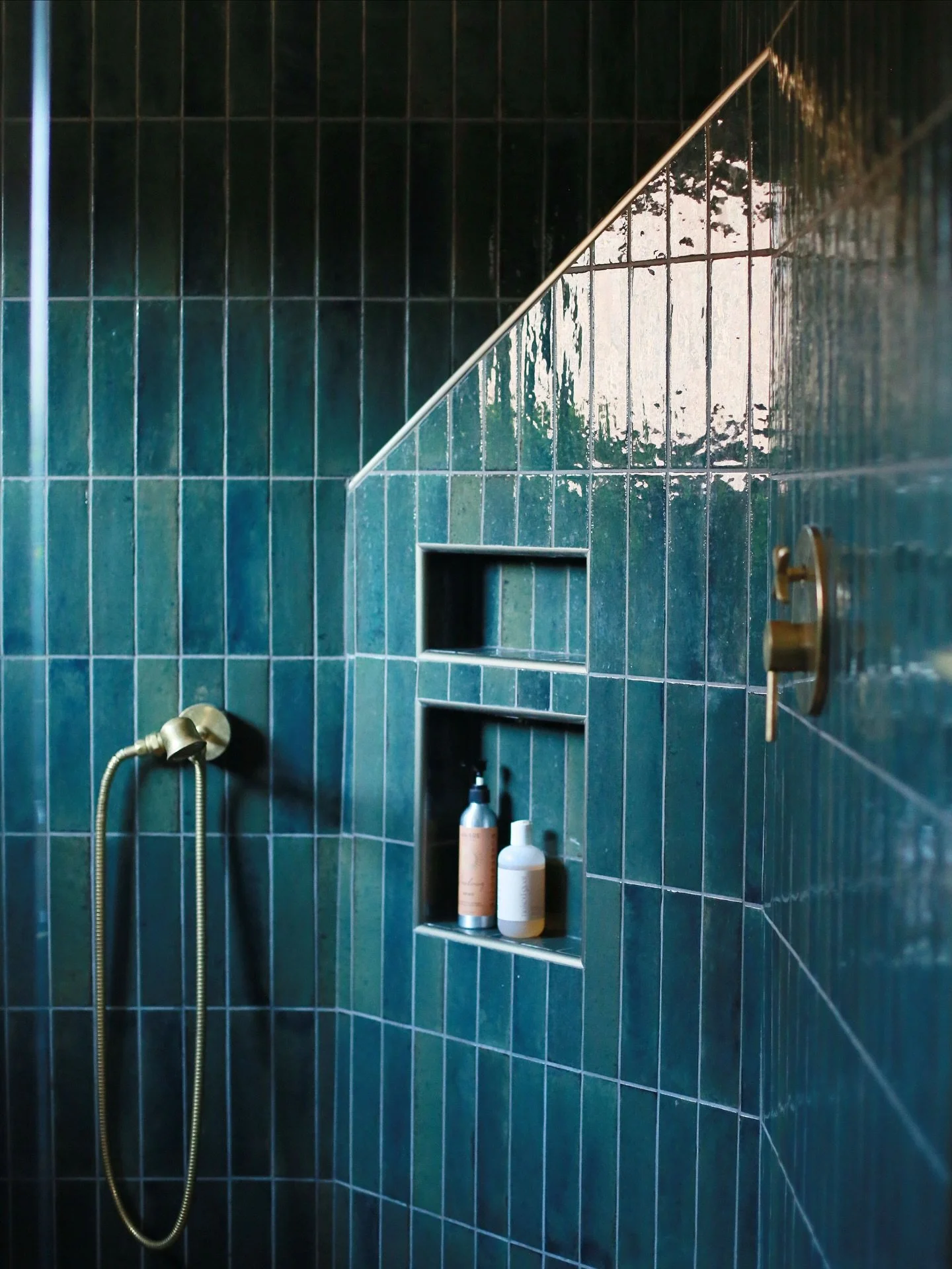 A modern shower with dark teal subway tiles, a built-in niche holding bottles of shampoo and body wash, a wall-mounted brass shower handle, and a brass handheld showerhead with a flexible hose.