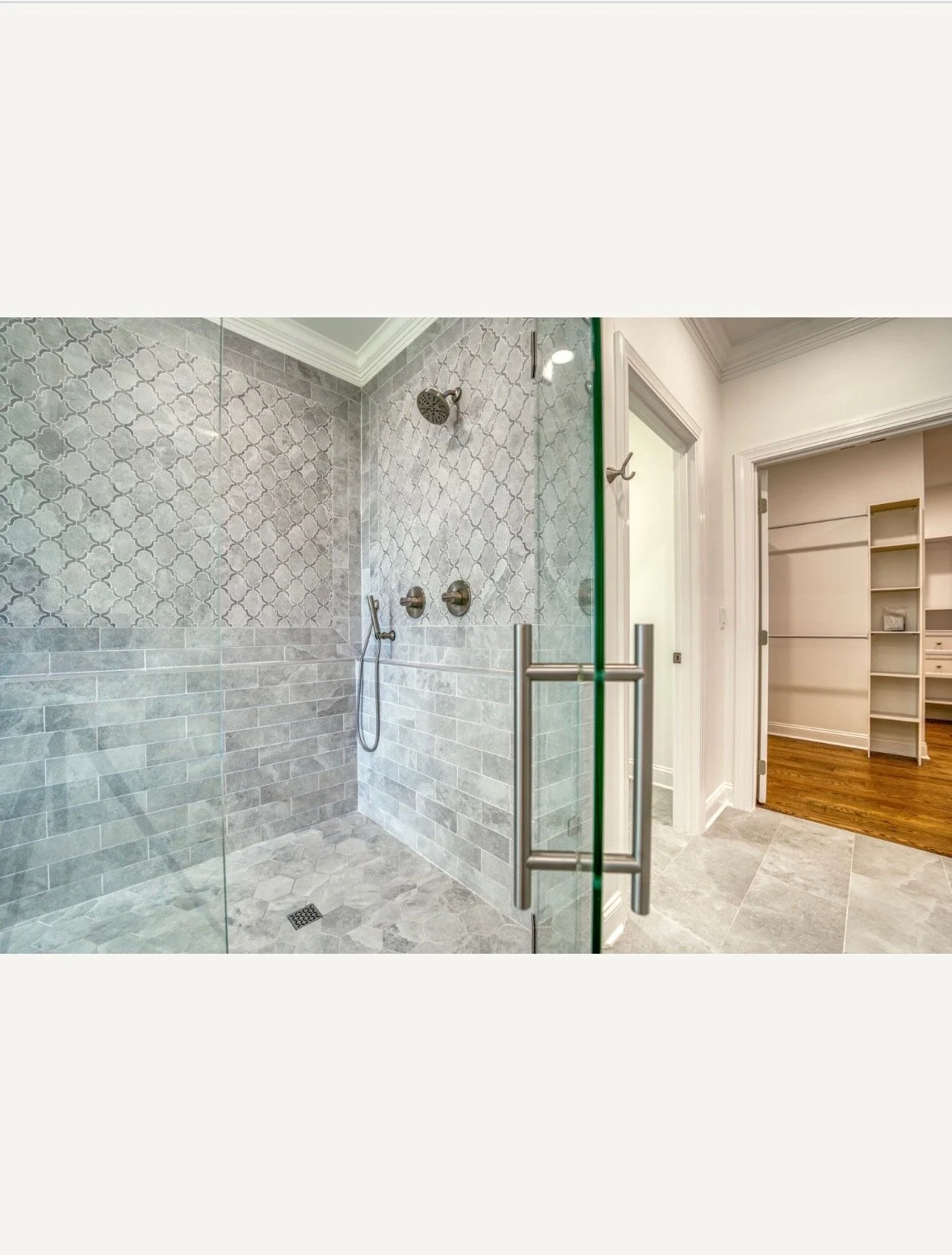 Glass-enclosed walk-in shower with patterned wall tiles, two showerheads, and a handheld shower in a modern bathroom with hardwood floor.