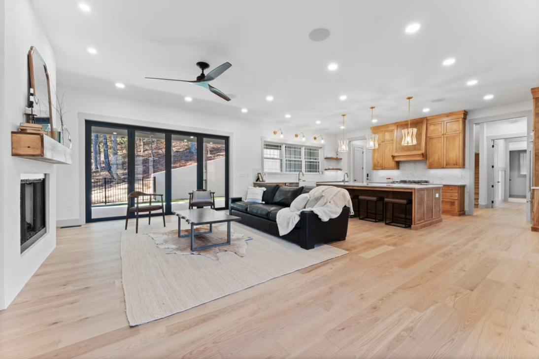 Open-concept living room and kitchen with hardwood floors, a black leather sofa, a fireplace, large sliding glass doors, and wooden kitchen cabinets with pendant lights.