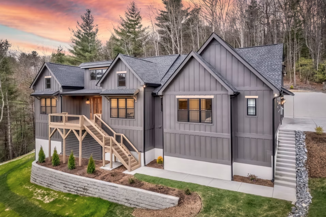 Modern gray house with multiple gabled roofs, a wooden staircase leading to the front porch, and landscaped yard with small trees, set against a sunset and wooded background.